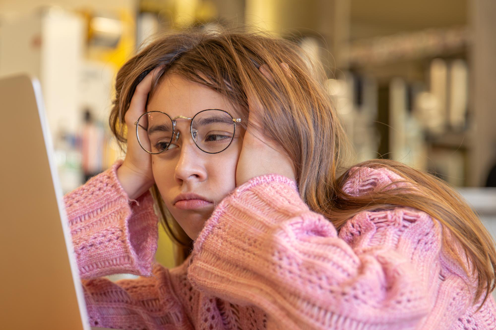 A young person wearing glasses and a pink sweater rests their head in their hands, looking frustrated or bored while staring at a laptop—possibly searching for an ADHD service online.