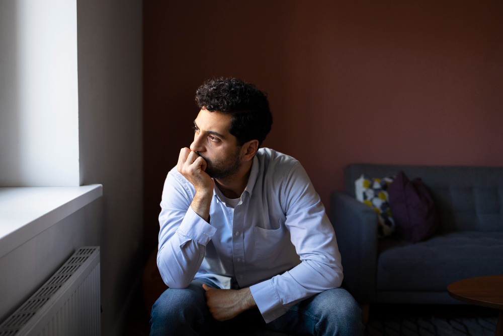 A man in a white shirt sits on a gray couch, resting his chin on his hand and looking thoughtfully out a window—capturing the quiet reflection often seen in those navigating Anxiety Disorders, supported by Newinsights Behavioral Health.