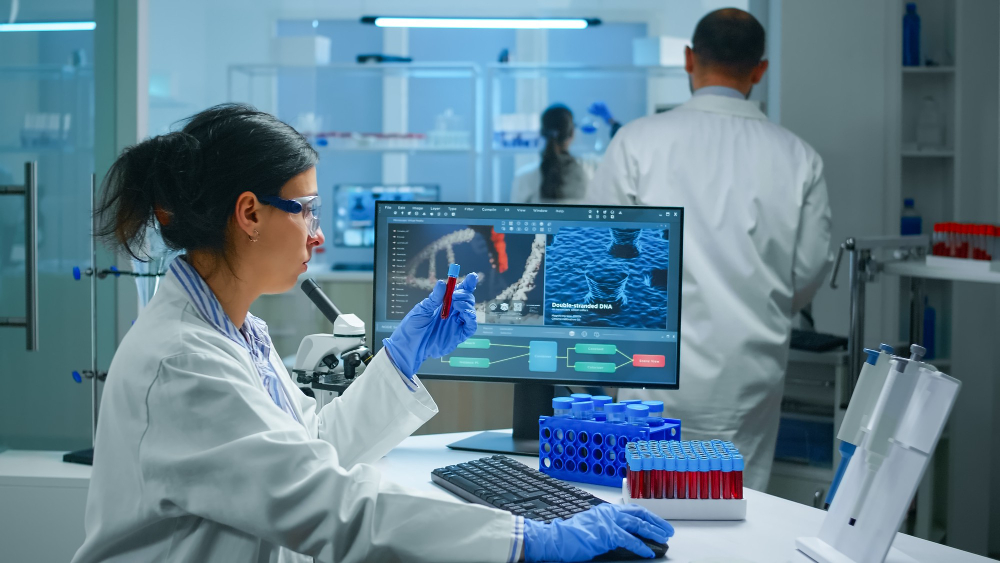 A female scientist in a lab coat and safety glasses examines a test tube at a desk with a computer displaying GeneSight® Testing results. Another scientist works in the background surrounded by lab equipment and test tubes.
