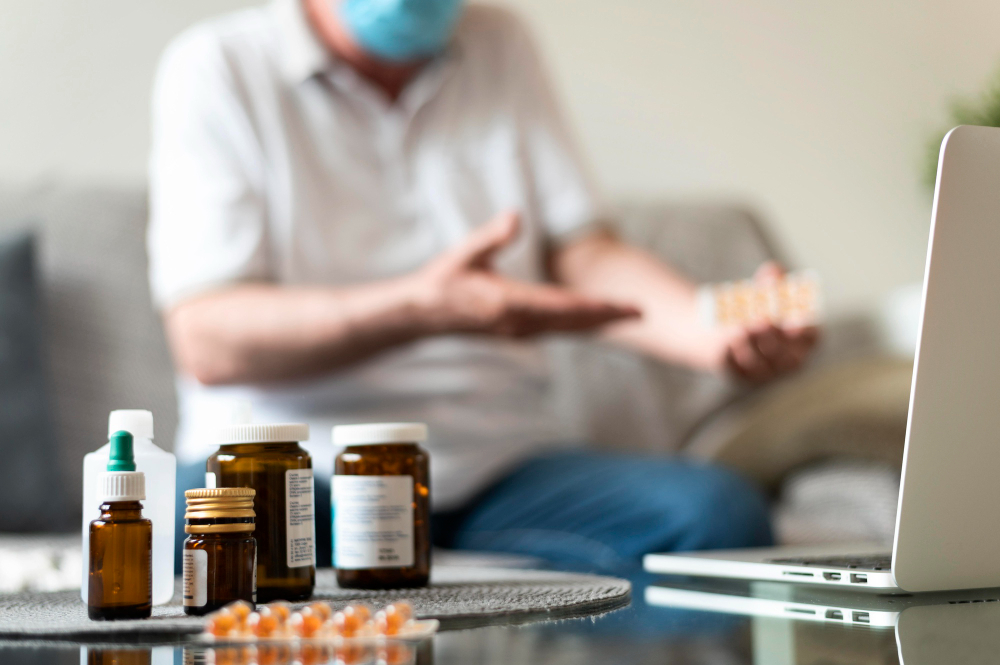 Several medicine bottles and pill packets are on a table in focus, while a person sits in the background on a couch, highlighting the need for Medication Management Services at New Insights Behavioral Health across Connecticut.
