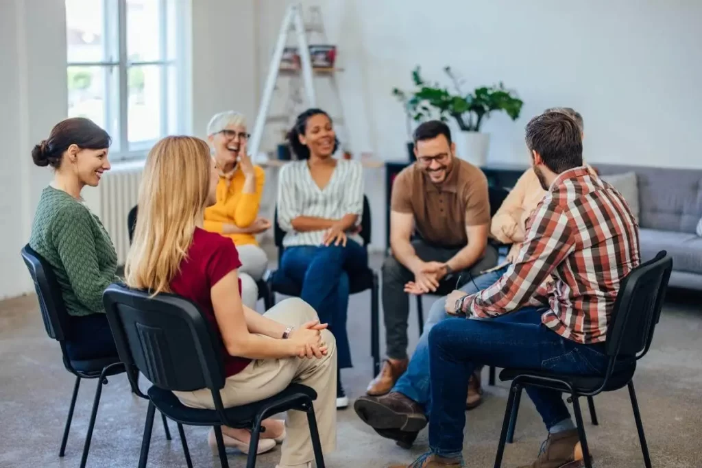 A group of seven adults sit in a circle on chairs in a bright room, engaged in lively conversation and laughter, suggesting a friendly support group or discussion meeting focused on trauma & stress-related disorders.